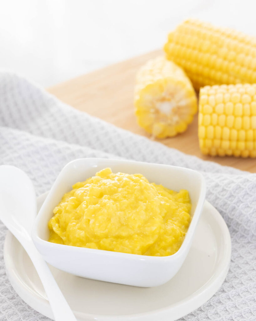 Sweet Corn Puree served for baby, with whole corn cobs in the background.