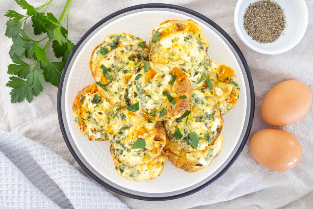 Pumpkin and Feta Egg Bites served on a platter, with ground black pepper, parsley and eggs around the plate.